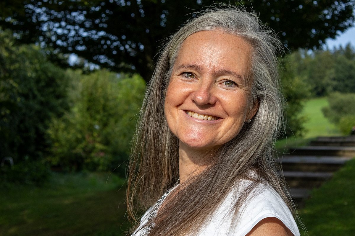 weibliche Person, halblange graubraune Haare, lächelt, im Hintergrund Natur